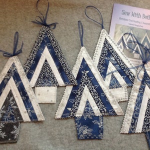 May include: A collection of handmade Christmas tree decorations. The triangular trees are crafted from blue and white patterned fabric, with a central white stripe. Each ornament has a blue ribbon. A booklet titled "Sew With Beth" is also visible.