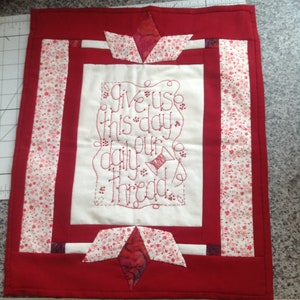 May include: A red and white quilted wall hanging with the embroidered text "Give us this day our daily thread." The design includes floral patterned fabric and a spool of thread.