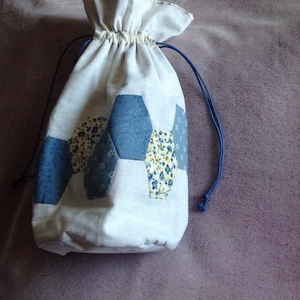 May include: A cream-coloured drawstring bag with a patchwork design of blue and floral patterned hexagons. The bag has a gathered top and blue drawstring cords.