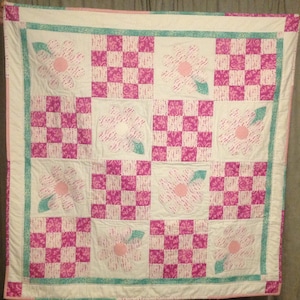 May include: A handmade quilt with a repeating pattern of pink and white flowers and checkered squares. The flowers have pink centres and dotted petals, with a teal leaf. The quilt is bordered with a teal stripe and white fabric.