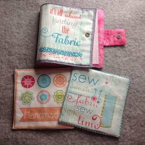 May include: A sewing kit with a light blue cover, featuring the text "It's all about finding the Fabric." The kit has a pink snap closure and two fabric squares with sewing-themed designs and text. One reads "Handmade."