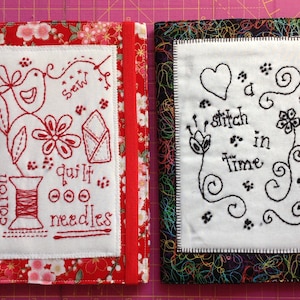 May include: Two fabric-covered journals. One has a red floral pattern with a red elastic band and a white square with red embroidery of a bird, flowers, and text. The other has a black and rainbow pattern with a green elastic band and a white square with black embroidery of a heart, flowers, and text.