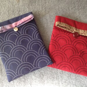 May include: Two fabric cases, one navy blue and one red, each with a stitched semi-circle pattern. The navy case has a light pink and white patterned flap, while the red case has a beige and brown patterned flap. Both have a button closure.