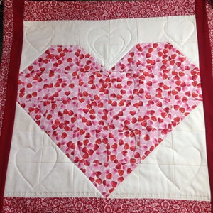 May include: A quilted table runner with a heart design. The heart is made of pink fabric with red hearts, set against a white background with heart-shaped quilting. The border is red with a floral pattern.