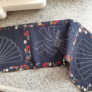 May include: Navy blue quilted fabric featuring white embroidered fan and bow motifs. The edges are trimmed with a floral pattern in red, brown, and green. Likely a decorative table runner or placemat.