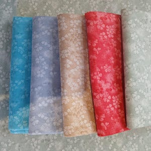 May include: Rolled fabric bundles in shades of blue, beige, and red, each decorated with a white floral pattern. The fabric is arranged on a light blue floral patterned background.