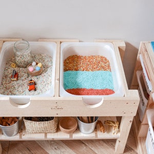 May include: A wooden play table with two white plastic trays filled with colourful rice. The table has a lower shelf with storage baskets and a small wooden toy. The table is labelled "Vacademy".