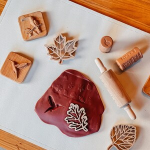 May include: A set of wooden stamps featuring various nature-themed designs, including leaves, birds, and a snail. The stamps are arranged on a white surface with a rolling pin and a red clay mat with impressions of the stamps.