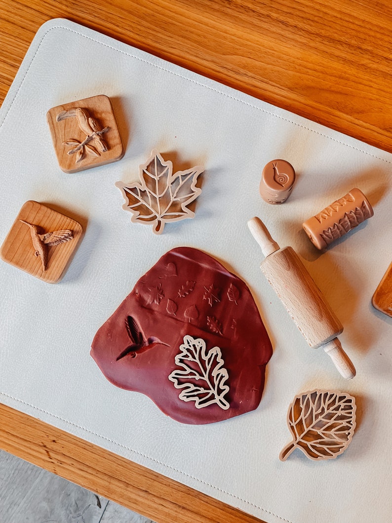 May include: A set of wooden stamps featuring various nature-themed designs, including leaves, birds, and a snail. The stamps are arranged on a white surface with a rolling pin and a red clay mat with impressions of the stamps.