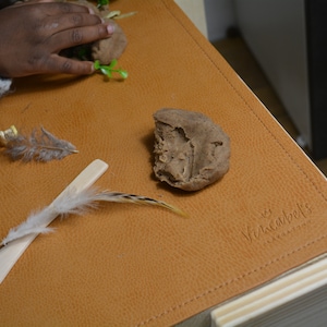 May include: A brown leather play mat with a wooden frame. The mat has a variety of items on it, including a small yellow rubber duck, feathers, a wooden spoon, and a lump of brown play dough. The mat has the text "Versarebels" printed on it.