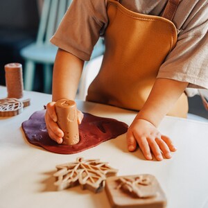 May include: A child uses a wooden leaf-shaped cookie cutter to press into red play dough on a white surface. Other wooden cookie cutters and a rolling pin are also visible.