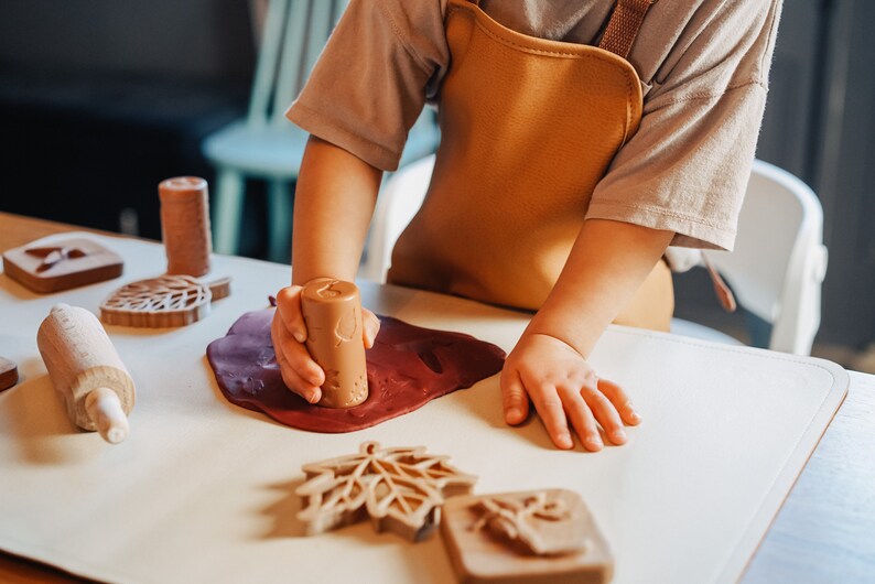 May include: A child uses a wooden leaf-shaped cookie cutter to press into red play dough on a white surface. Other wooden cookie cutters and a rolling pin are also visible.