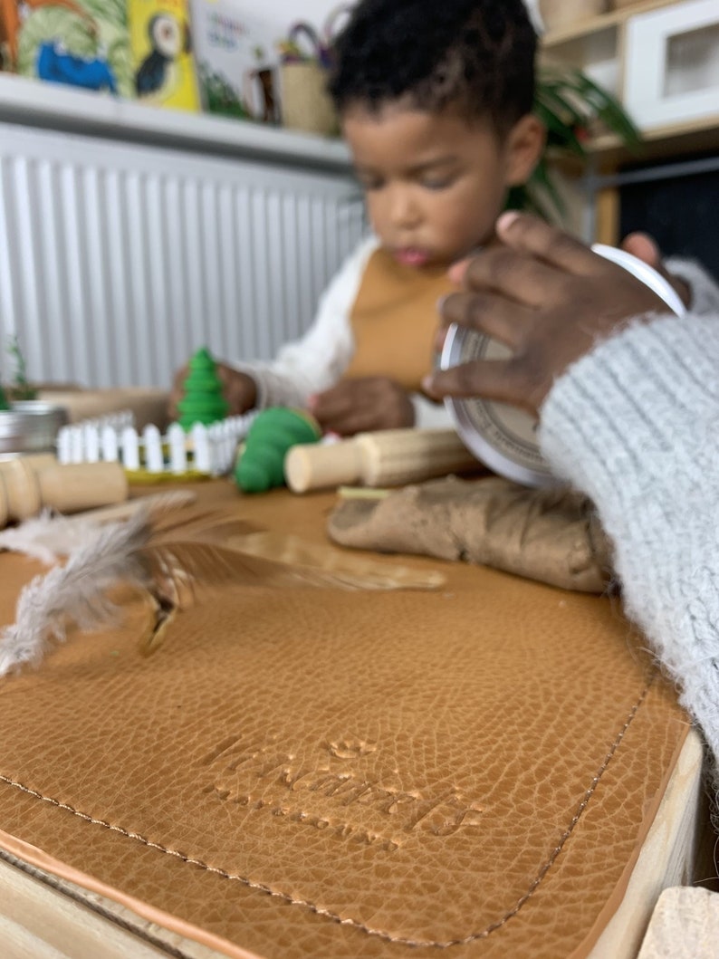 May include: A child is playing with play dough on a brown leather play mat with the words "The Playmat Co." embossed on it. The mat has a stitched edge.