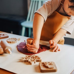 May include: A child wearing a brown apron uses a wooden stamp to press a leaf shape into red play dough.  The child is surrounded by other wooden stamps and a rolling pin on a white surface.