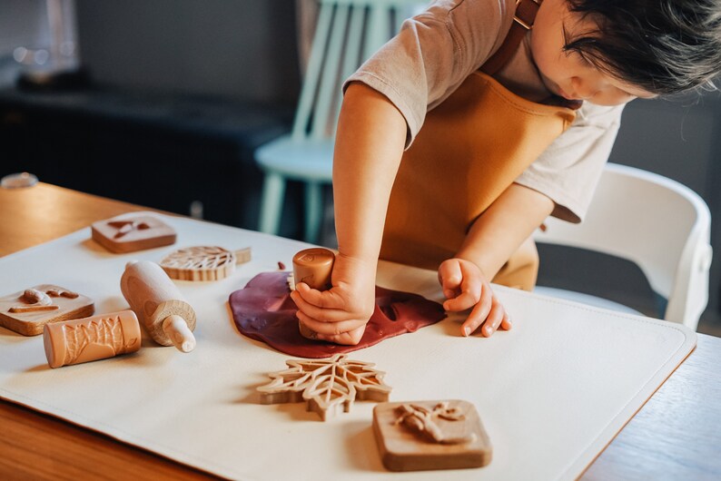 May include: A child wearing a brown apron uses a wooden stamp to press a leaf shape into red play dough.  The child is surrounded by other wooden stamps and a rolling pin on a white surface.