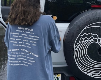 May include: A white Jeep with a spare tire cover featuring a black and white wave design. The spare tire cover is on the back of the vehicle. A person is standing next to the Jeep wearing a blue t-shirt with the text "Mighty God Friend of Sinners" and a list of words below it.
