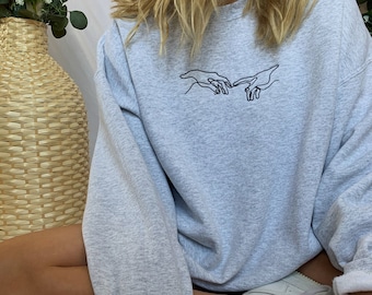 May include: A gray sweatshirt with a black embroidered design of two hands reaching towards each other.