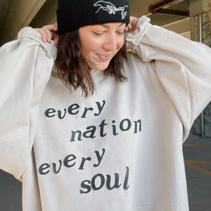 May include: A cream-colored hoodie with the text "every nation every soul" printed in black.