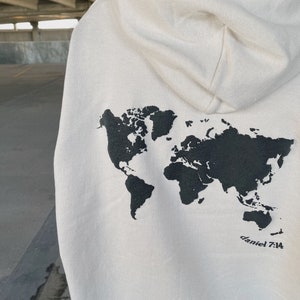 May include: A cream-colored hoodie with a black outline of the world map printed on the back. The text "daniel 7:14" is printed below the map.