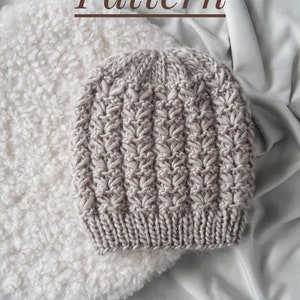 Peut inclure: Gros plan d'un bonnet tricoté de couleur gris clair avec un motif en côtes. Le bonnet est posé sur une surface blanche et moelleuse.