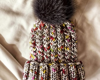 May include: A knitted hat with a large, dark gray fur pom-pom. The hat is a mix of white, gray, and colorful flecks of red, yellow, and pink. The words "Knitting Pattern" are at the top.