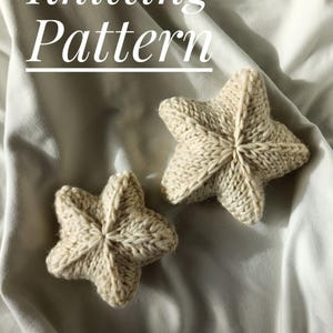 May include: Two knitted beige star-shaped ornaments on a white fabric background. The text "Knitting Pattern" is at the top of the image.