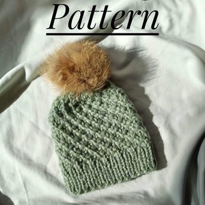 May include: A knitted beanie in a soft green hue, topped with a fluffy brown pom-pom. The hat features a textured stitch pattern and the words "Knitting Pattern" in black script above it. The hat is displayed on a white fabric background.