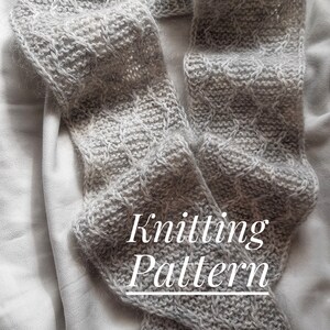 May include: A gray knitted scarf with a cable stitch pattern. The scarf is folded in half and laid on a white fabric background. The text "Knitting Pattern" is written in white on the scarf.