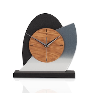 Könnte beinhalten: Eine moderne Tischuhr mit einem Holzgesicht und silbernen Zeigern. Die Uhr ist vor einem schwarzen und silbernen geometrischen Design angeordnet und auf einem schwarzen Sockel montiert. Das Zifferblatt der Uhr ist aus Holz und die Zeiger sind silber.