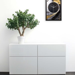 May include: A black and gold abstract wall clock with a white background. The clock is framed in black and is hanging on a white wall. A small potted plant is in front of a white dresser with two drawers.
