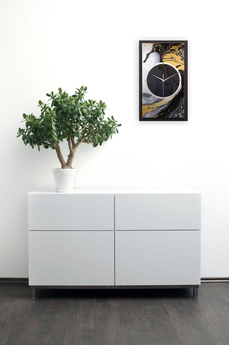May include: A black and gold abstract wall clock with a white background. The clock is framed in black and is hanging on a white wall. A small potted plant is in front of a white dresser with two drawers.