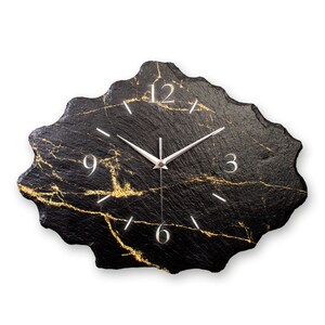 Könnte beinhalten: Schwarze und goldene Uhr mit einer einzigartigen, unregelmäßigen Form. Das Zifferblatt der Uhr hat römische Ziffern und einen silberfarbenen Stunden- und Minutenzeiger.