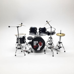 May include: Miniature black drum set with silver hardware. The bass drum features the text "GREEN DAY American Idiot" and a hand holding a red grenade. Includes cymbals, drumsticks, and a small stool.