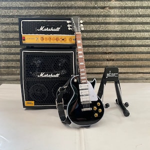 Könnte beinhalten: Schwarze E-Gitarre mit weißem Schlagbrett und goldenen Hardware, ein schwarzer Verstärker mit dem Wort "Marshall" darauf und ein schwarzer Gitarrenständer. Der Verstärker trägt die Aufschrift "JCM 900" und "1960A".