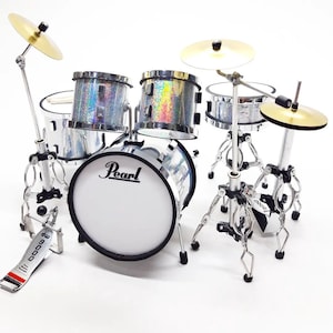 Könnte beinhalten: Ein Miniatur-Schlagzeug mit silbernem Glitzer-Finish. Das Set beinhaltet eine Bassdrum mit weißem Pearl-Logo, zwei Toms, eine Snare Drum und zwei Becken. Die Trommeln sind auf Ständern mit verstellbaren Beinen montiert.