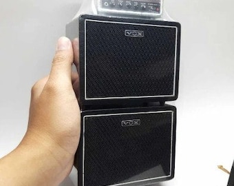 May include: A black Vox guitar amplifier with two speakers. The top speaker is smaller and silver with the Vox logo. The bottom speaker is larger and black with the Vox logo.