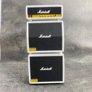 May include: A stack of three white Marshall amplifiers. The top amplifier has a black face with the Marshall logo and gold accents. The middle and bottom amplifiers have black speaker covers with the Marshall logo. Each has a small yellow label that reads "JCM 800 LEAD - 1960".