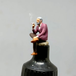 May include: A miniature figurine of a man with grey hair and a beard, wearing a burgundy jacket and brown trousers, sitting on a small rock and holding a cigarette. Smoke is coming from the cigarette.