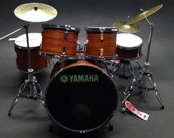 May include: Miniature drum set with a dark wood finish. The set includes a bass drum with the Yamaha logo, several toms, cymbals, and a kick pedal. The cymbals are gold-colored, and the drumheads are white. The set is on a black surface.
