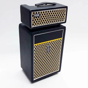 Op de afbeelding: Een zwarte en gouden Vox gitaarversterker stack. De versterker heeft een getextureerde gouden luidsprekergrille, een zwarte handgreep en het Vox-logo. De stack bestaat uit een top en een cabinet.
