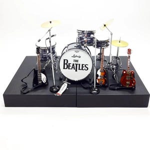 May include: Miniature drum kit with guitars. The bass drum features "The Beatles" logo. The set includes a bass drum, snare drums, cymbals, microphones, and three guitars. The set is displayed on a black platform.
