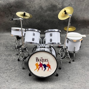 May include: Miniature white drum set with silver hardware and gold cymbals. The bass drum features "The Beatles" logo with colorful silhouettes of the band members. Includes drumsticks.