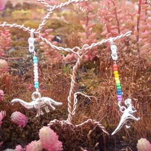 May include: Silver dinosaur earrings with colorful beaded accents. One earring features a dinosaur with a rainbow of beads, the other with pastel beads. The earrings hang from a copper wire structure, set against a backdrop of pink and green plants.
