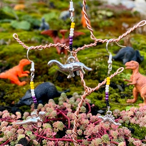 May include: Earrings featuring silver dinosaur pendants and non-binary pride flag beadwork. The earrings are suspended from a copper wire sculpture, with toy dinosaurs visible in the background. The setting includes moss and succulent plants.