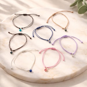 May include: A selection of adjustable string bracelets in different colours, each with a small charm. Colours include brown, tan, blue, purple, pink, and white. Charms are various shapes and colours, including turquoise, green, and clear.