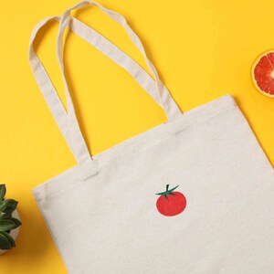 May include: A white canvas tote bag with a red embroidered tomato on a yellow background. There are also three orange halves of citrus fruit on the background.