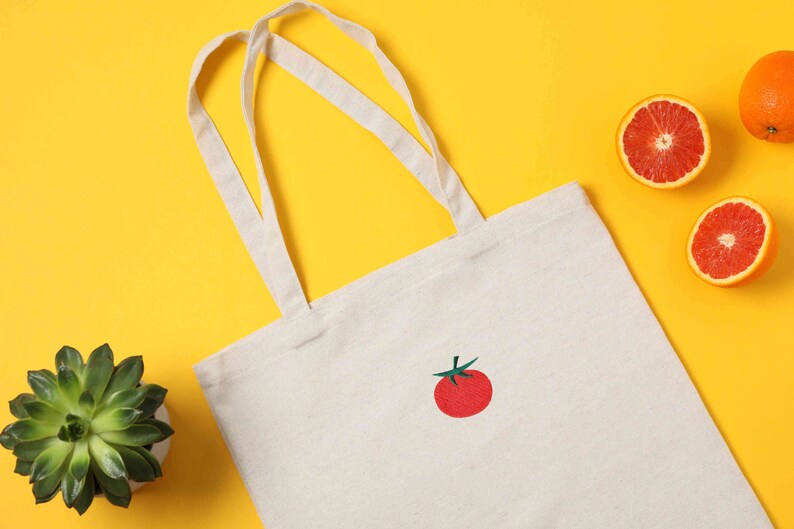 May include: A white canvas tote bag with a red embroidered tomato on a yellow background. There are also three orange halves of citrus fruit on the background.