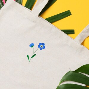 May include: Cream-colored tote bag with embroidered blue forget-me-not flowers. The flowers have yellow centers and green stems and leaves. The bag has two handles and is made of a natural fabric.