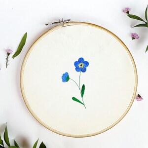 May include: An embroidery hoop with a cream-colored fabric featuring a blue forget-me-not flower design. The flower has a yellow center and green stem and leaves. The wooden hoop is round and has a metal clasp.