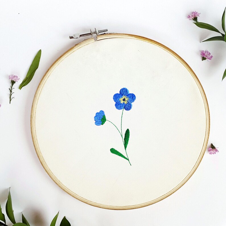 May include: An embroidery hoop with a cream-colored fabric featuring a blue forget-me-not flower design. The flower has a yellow center and green stem and leaves. The wooden hoop is round and has a metal clasp.
